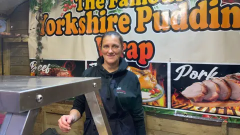 Kate Andrews stands in her Yorkshire Pudding wrap stall at swindon Christmas market, she is wearing a black hoodie and apron and in front of an advertising poster showing what the stall sells