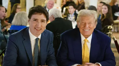 Justin Trudeau and Donald Trump, both in blue suits, sit shoulder to shoulder with people seen in a crowd behind them 