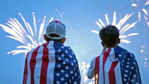Two boys watch a fireworks display during a celebration of the US Army's 250th anniversary on Flag Day, June 14, 2025 in Washington DC. They are draped in USA flag and the sky in front of them is alight with fireworks