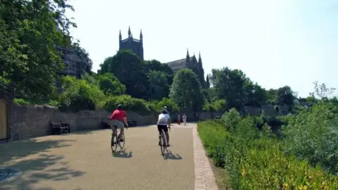 Two people riding on bicycles along a pathway that is surrounded by green grass, bushes and trees. In the background is the tower of a church