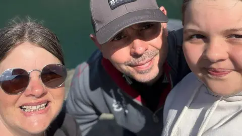 Jenni Whitmore A selfie on Jenni with her husband Gareth and young daughter Carys. Jenni has her hair tied back an is wearing sunglasses. Her husband and daughter are wearing grey hoodies. Her husband is wearing a grey baseball cap.