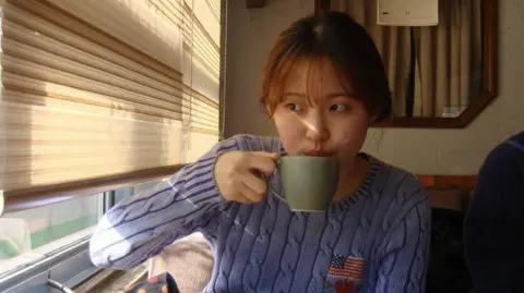 Soomin Yi Soomin Yi sipping on a cup of tea while glancing towards a window to her right