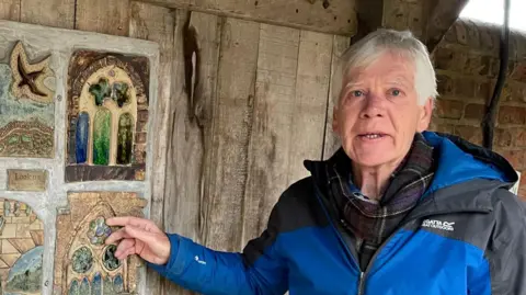 BBC A man with grey hair in a blue coat pointing at decorated tiles on a wooden door. The tiles show small raised pottery artwork of windows and birds.