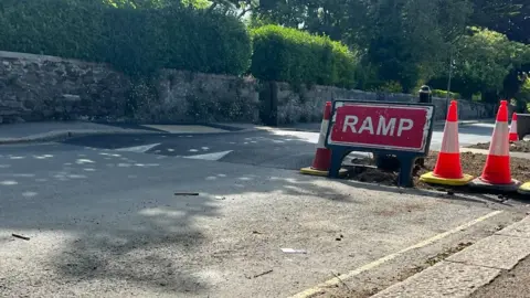 LDRS A table-top speed hump on Malpas Road in Truro. Traffic cones and a red warning sign saying ramp and on the right-hand side. A stone wall, hedges and trees are on the left-hand side of the road.