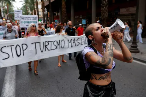 Reuters A demonstrator with tattoos uses a megaphone as people protest against mass tourism on a street in Alicante, Spain, July 13, 2024. The banner reads "Stop turismo masivo" ("Stop mass tourism")