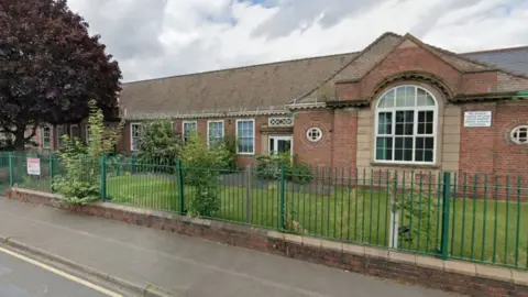 The school looks to be on one level and has large glass windows and built with red bricks and a green fence in front of it.