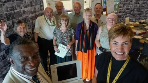 Ten people pose around an old computer, looking up and smiling at a camera held above. They are in a room with an exhibition of old black and white photos on the wall and tables on the right set for lunch. There are five men and five women.