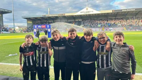 LADY BAY PRIMARY SCHOOL 7 boys stood in a line pitch side at Meadow Lane, Notts County. Some are in Notts County kit.