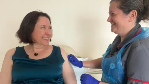 Clare wears a blue, sleeveless top and sits next to a nurse who is rubbing Clare's left arm and is wearing purple medical gloves, ready to vaccinate her