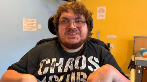 Isaac Wide is wearing a black t-shirt which reads "chaos club" and is sitting in a wheelchair next to a bed. He has a brown cropped beard, brown curly hair and is wearing glasses. The walls behind him are painted a pale blue and yellow.
