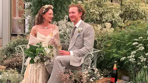A couple are posing as newlyweds at a wedding reception. They are sitting on a garden bench with glasses of champagne, and behind them the garden is planted with white flowers. There is a bottle of champagne to one side. The woman is holding a bouquet.