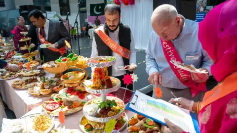 Supplied/BSCMS A long table full of colourful food with people stood helping themselves. one person has a clipboard.