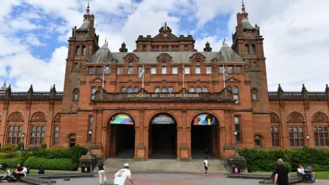 Getty Images Historic red‑sandstone building with ornate towers and archways, displaying the signage ‘Kelvingrove Art Gallery and Museum,’ with people walking and sitting in the open area in front