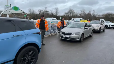 A row of cars next to crates of bottled water on the left hand side.