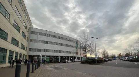 The exterior of Great Western Hospital on a cloudy winter's day. There is a large beige coloured building with a modern white cladded one with lots of windows, with a large car park out the front. People can be seen walking down the path.