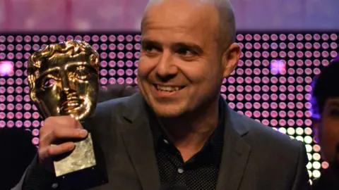 Oli Hyatt wearing a suit and stood on a stage holding a BAFTA award.