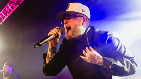 Thom Rylance is wearing a white cap, orange tinged glasses and black jacket. He singing into a microphone on stage.