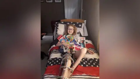 A young boy lying on a single bed with a leg in a frame. He is smiling at the camera. There are magazines and books over his chest.