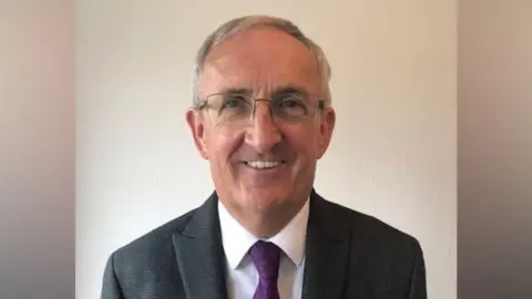 A man with short grey hair and glasses smiles at the camera. He is wearing a dark grey jacket, white shirt and purple tie. 