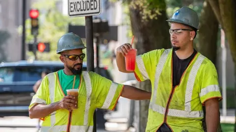 AFP via Getty Images Two construction workers wearing hi-vis jackets and helmets take a drinks break in Everett, Massachusetts.