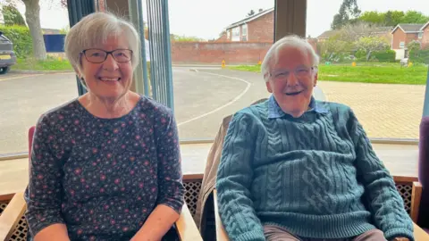 Sandra and John Bishop are pictured sitting in chairs in front of a large window. A housing estate and a green area can be seen out the window. Both Sandra and John are smiling. Sandra has short, white hair and is wearing a dark purple patterned top. John also has white hair and is wearing a light blue jumper and grey trousers.
