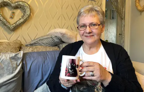 A woman with short blonde hair and glasses sites in a smart sitting room which has golden-coloured wallpaper and gold heart decorations. She is holding a mug with a picture of a cat and wearing a T-shirt with a similar picture.