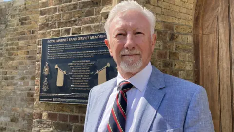 Derek Lindars is standing in front of a memorial plaque. He is wearing a pale blue jacket, white shirt and striped red, black and yellow tie