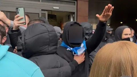 Crowd gathered outside a building entrance, with several people in dark hooded jackets and one individual raising an arm while holding a scarf around the neck, others recording with smartphones.