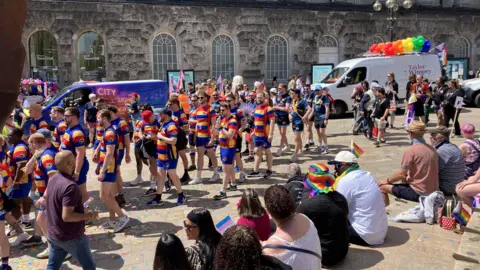 dozens of people taking part in a parade. the people in the picture are dresses in blue short with blue, red and yellow striped tops. There is a van in the background with rainbow balloons strapped to its roof. People are sitting on nearby steps with rainbow flags