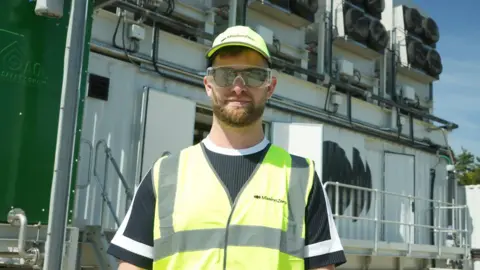 Jamie Niblock/BBC Nicholas Chadwick standing outside the air capture plant, wearing sunglasses with protective goggles over the top. He wears a black and white t-shirt and a high vis vest and baseball cap.
