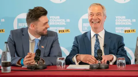 Two men sit next to each other with a blue banner in the background that advertises Reform UK. They are both wearing suits