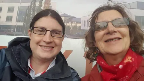 Sarah Edgeworth Sarah Edgeworth wearing a black hooded raincoat and black rectangular glasses, with her dark hair pulled back into a ponytail with a middle part. She is smiling for a selfie next to her mum Sue, who is wearing a coral jacket, a red scarf and rectangular sunglasses.
