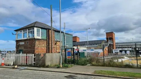 Geographic/Chris Morgan Newsham signal box is a square redbrick building and behind it is a footbridge crossing the line.