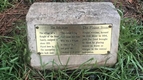 A gold plaque affixed to a small stone in the grass, detailing the origin of the Plague Stone and The Hob Stone. Under the Hob Stone entry it reads: 'The effigy of a Knight of the Roos family, sculpted in about about 1315.