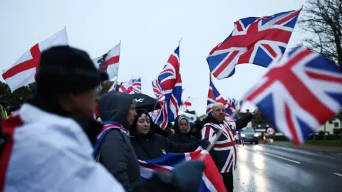 Reuters People waving Union Jacks and St George's flags by the side of the road in wet conditions. Some people have hoods and umbrellas up.