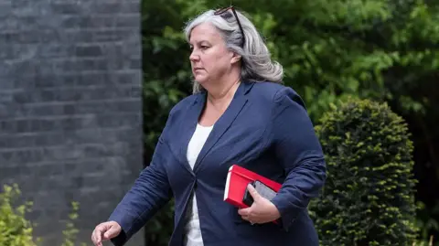 Getty Images Secretary of State for Transport Heidi Alexander arrives in Downing Street to attend the weekly Cabinet meeting in London on 28 April 2026. She is wearing a white top and blue jacket and has a red notebook and phone in her hand, with sunglasses in her hair.