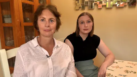 Susan and Anna - Susan sits on the left and wears a white shirt. Sh4e has brown hair and a neutral expression.