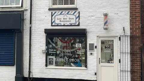 Image shows the outside of Lads and Dads Barbershop in Loughborough. The bricks on the building are white and so is the door. The window is in the middle and has tinsel dangling down it from the inside. There is also writing on both sides of the window saying 'walk-ins welcome,' and in the middle towards the bottom of the window it says 'Gentleman's Barber Shop' in large white writing. The logo is also above the window and says Lads and Dads Barbershop in caps on a white sign with blue, red and white stripes. 