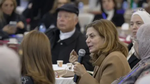 Kabawat speaking into a microphone at a conference in Idlib. She is seated at a table, with two women in headscarves on the right of the picture, and a man in a dark cap and a woman with long brown hair on the other side.