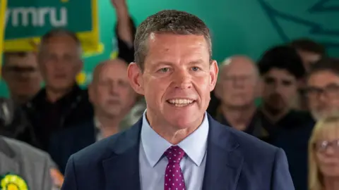 Leader of Plaid Cymru Rhun ap Iorwerth speaking after Plaid Cymru's Senned election victory. He is wearing a navy-blue blazer, light blue shirt, and a purple polkadot tie. He is smiling and looking at the camera. People can be seen stood behind him.