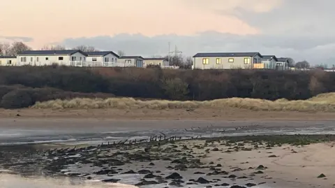 A row of metal poles can be seen emerging from the river bed across an entire estuary. On the opposite bank there are caravans.