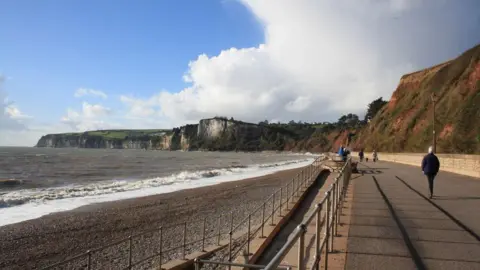 John Stephen The west walk at Seaton, with the shore on the left-hand side of the image and a large path on the right-hand side with metal fencing separating the two. In the distance there are large white cliffs making up headlands and next to the path there are red cliffs with a bit of grass covering them.
