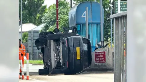 Paul Lynch A black Range Rover flipped onto its right hand side next to a large blue toll booth on Tamar Bridge. To the right of it is a red and white sign saying 'motorcycles and cycles must stop at toll booth'. To the left is a man in an orange hi-vis.