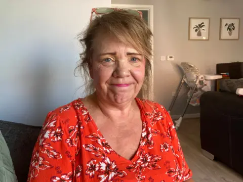 A woman in a red floral top smiling at the camera. She has blue eyes, brown hair down to her shoulders and glasses that are sat on the top of her head. In the background you see a baby high chair and two photos of plants on the wall