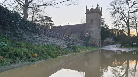 Stone wall leading from church beside road submerged in brown floodwater.