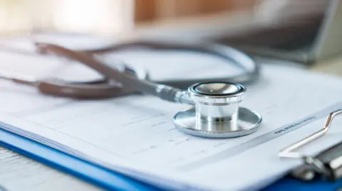 A stethoscope in silver metal is on top of a clipboard which has a blue plastic back and paper attached to it. The background is blurred but a laptop can be seen as well as the tube of the stethoscope out of focus.