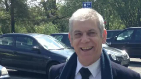 Thames Valley Police/Contributed John Jones: A grey haired man smiling. He is wearing a white shirt. blue tie, blue woollen scarf and blue coat. Four dark-coloured cars are visible in the background.