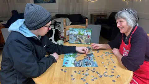 King's Arm Project Max, wearing a dark hat and glasses, with a blue hoodie, dark coat, sitting at a wooden table, making a jigsaw puzzle with Bernice, a woman, with short grey hair, with glasses on her head, wearing a purple top and red apron. Bernice is smiling and looking at Max. 