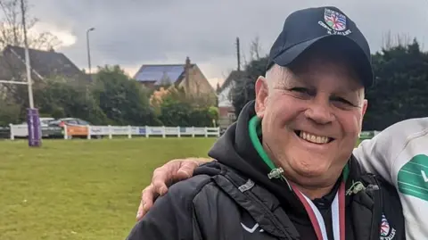 Chew Valley RFC Steve Worrall smiling, wearing a Chew Valley RFC cap and jacket, while standing on a pitch. Someone has his arm around Mr Worrall's shoulders