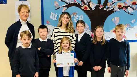 A group of six school children dressed in school uniforms consisting of blue jumpers, grey trousers and white polo shirts smile into the camera as they hold a healthy school certificate. The group are joined by two female adults who are also smiling. They are all standing in front of a mural within a school depicting birds on a tree which is covered in snow.